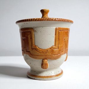 Puede incluir: Un tarro de cerámica con tapa. El cuerpo crema presenta diseños geométricos marrones. El tarro tiene una base con pie y un borde texturizado. La tapa tiene un pequeño pomo. El diseño es rústico y artesanal. Altura aproximada 15 cm.
