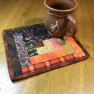 May include: A brown ceramic mug with white leaf designs sits on a handmade, rectangular quilted coaster. The coaster features a patchwork design with various fabrics in shades of brown, orange, and green. The mug and coaster are on a wooden surface.