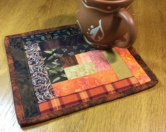 May include: A brown ceramic mug with white leaf designs sits on a handmade, rectangular quilted coaster. The coaster features a patchwork design with various fabrics in shades of brown, orange, and green. The mug and coaster are on a wooden surface.