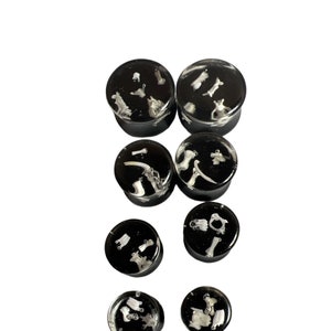 May include: Set of seven black resin ear gauges with white bone fragments embedded inside. The gauges are various sizes and are arranged in a staggered pattern.