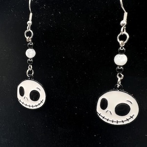 May include: Dangle earrings featuring a black and white Jack Skellington face design. The earrings have a silver-tone hook and chain with black and white beads. The earrings are a fun accessory for fans of the movie.