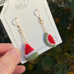 May include: Handmade watermelon slice earrings. Each earring features a crocheted watermelon slice in red, white, and green, with tiny black seed details. The earrings hang from gold-colored chains and hooks. The tag reads "We All Love Crafts Handmade".