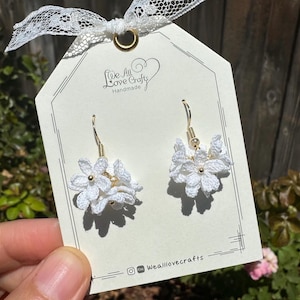 May include: White crochet flower earrings with gold-colored hooks and accents. The earrings are displayed on a tag that reads "We All Love Crafts Handmade". The tag is tied with a white lace ribbon.