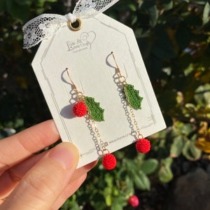 May include: Handmade crochet earrings featuring red berry-like spheres and green leaf accents. The earrings hang from gold-colored hooks and chains. The tag reads "We All Love Crafts Handmade".