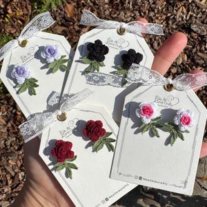 May include: Handmade crochet flower earrings in various colors, including purple, black, red, and pink, displayed on white tags with lace ribbon. Each tag has the text "We All Love Crafts Handmade" and the Instagram handle "Wealllovecrafts".
