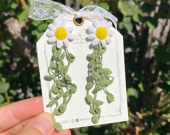 May include: Handmade daisy earrings with white petals and yellow centers, accented by green vine-like strands. The earrings are attached to a tag with the text "We All Love Crafts" and a white lace ribbon. The earrings are a unique floral accessory.