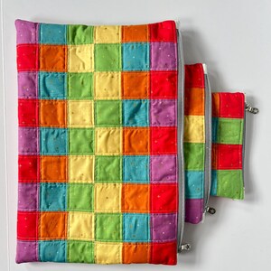 May include: Three zippered pouches made of quilted fabric with a rainbow patchwork design. The pouches are lined up in order of size, with the largest pouch on the bottom and the smallest pouch on top.