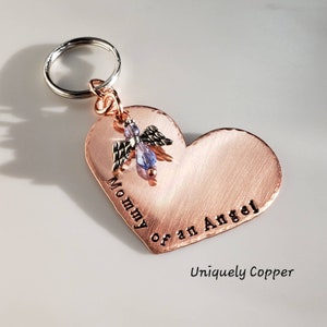 May include: Copper heart-shaped keychain with the text "Mommy of an Angel" engraved on it. A small angel charm with a purple crystal is attached to the top of the heart. The keychain is attached to a silver key ring.