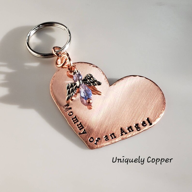 May include: Copper heart-shaped keychain with the text "Mommy of an Angel" engraved on it. A small angel charm with a purple crystal is attached to the top of the heart. The keychain is attached to a silver key ring.