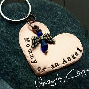 May include: Copper heart-shaped keychain with a blue angel charm and the text "Mommy of an Angel" engraved on it. The keychain is attached to a silver key ring. The text "Uniquely Copper" is written in cursive at the bottom of the image.