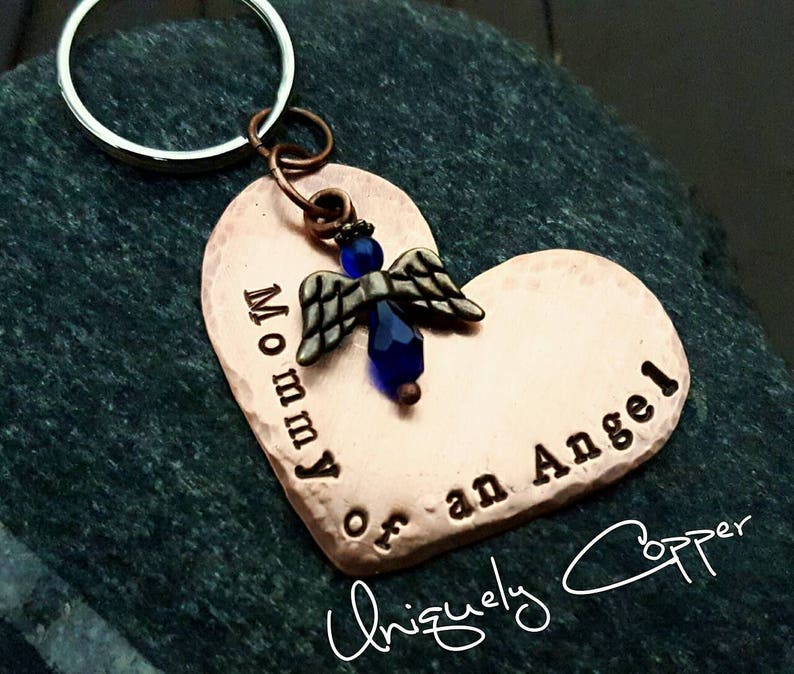 May include: Copper heart-shaped keychain with a blue angel charm and the text "Mommy of an Angel" engraved on it. The keychain is attached to a silver key ring. The text "Uniquely Copper" is written in cursive at the bottom of the image.