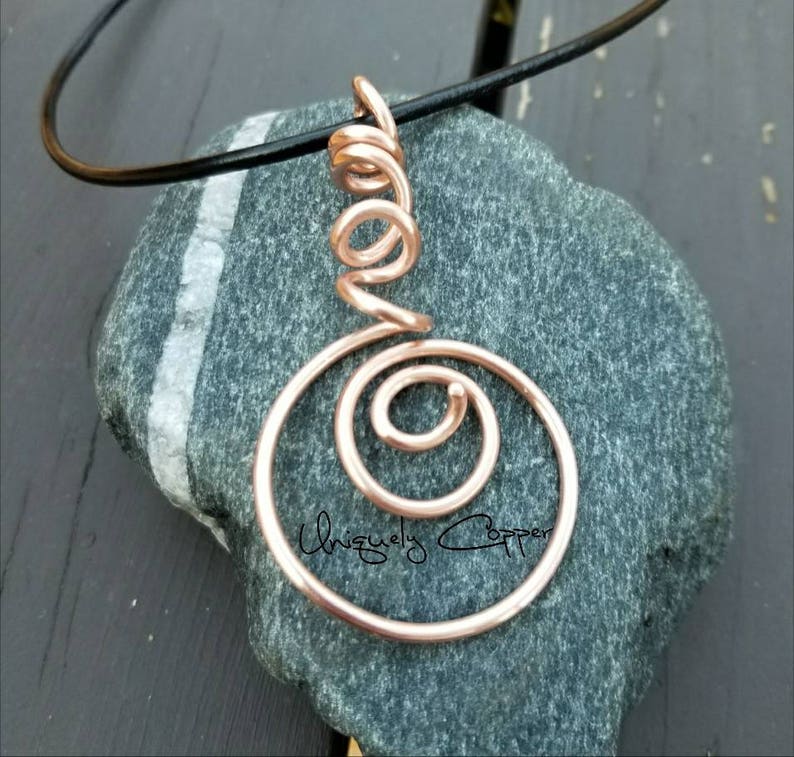 May include: A rose gold wire pendant necklace with a spiral design. The pendant is hanging from a black leather cord.