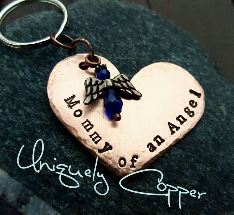 May include: Copper heart-shaped keychain with a small angel charm and blue crystal bead. The keychain is engraved with the text "Mommy of an Angel".