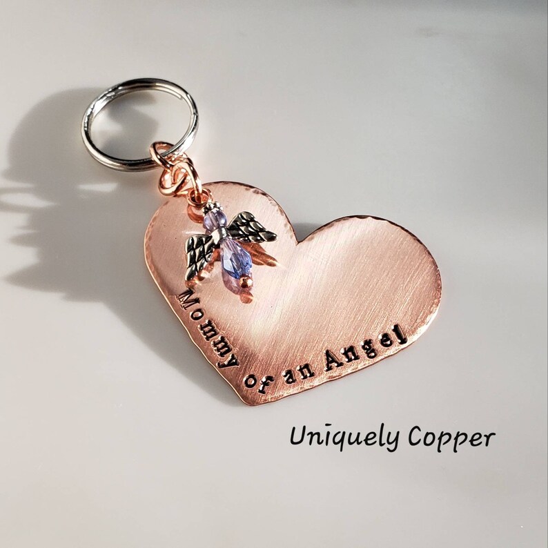 May include: A copper heart-shaped keychain with a small angel charm and the words "Mommy of an Angel" engraved on it. The keychain is attached to a silver key ring. The words "Uniquely Copper" are printed below the heart.