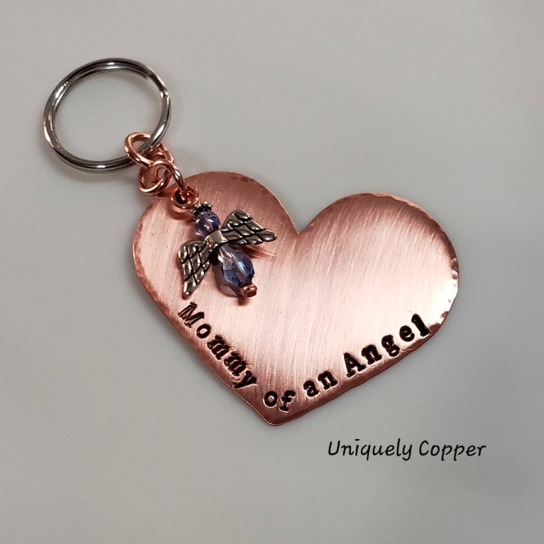 May include: A copper heart-shaped keychain with the words "Mommy of an Angel" engraved on it. A small angel charm with a blue crystal is attached to the heart. The keychain is attached to a silver key ring. The words "Uniquely Copper" are printed below the heart.