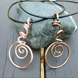 May include: A copper wire necklace with two spiral pendant charms. The necklace is on a black leather cord and is resting on a gray rock.