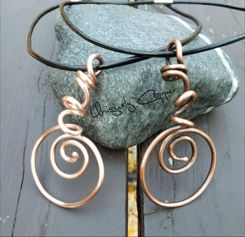 May include: A copper wire necklace with two spiral pendant charms. The necklace is on a black leather cord and is resting on a gray rock.