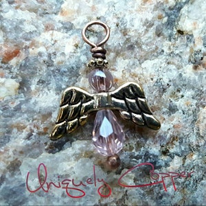 May include: A small copper angel charm with pink glass beads and gold wings. The charm is on a rocky surface.