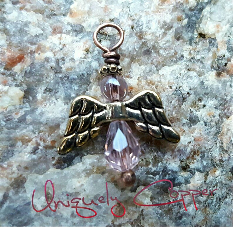 May include: A small copper angel charm with pink glass beads and gold wings. The charm is on a rocky surface.