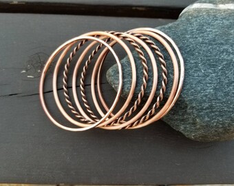 May include: A set of eight copper bangles, some with a twisted design, arranged in a circle on a dark gray surface.
