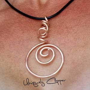 May include: A copper necklace with a spiral pendant. The pendant is made of wire that is twisted into a spiral shape. The necklace is on a black cord.