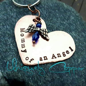 May include: Copper heart-shaped keychain with the words "Mommy of an Angel" engraved on it. A small angel charm with blue wings and a blue crystal bead hangs from the top of the heart.
