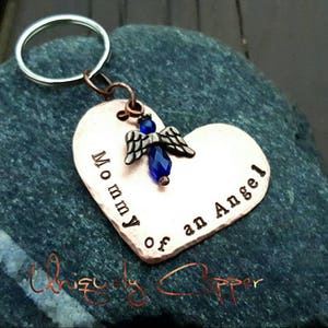 May include: Copper heart-shaped keychain with the words "Mommy of an Angel" engraved on it. A small blue angel charm with a blue crystal is attached to the top of the heart.