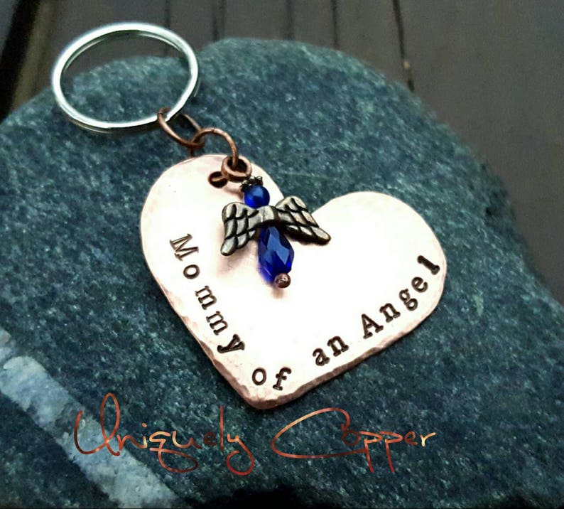 May include: Copper heart-shaped keychain with the words "Mommy of an Angel" engraved on it. A small blue angel charm with a blue crystal is attached to the top of the heart.