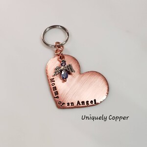 May include: Copper heart-shaped keychain with the text "Mommy of an Angel" engraved on it. A small silver angel charm with a blue crystal is attached to the heart. The keychain is attached to a silver key ring. The text "Uniquely Copper" is printed below the heart.