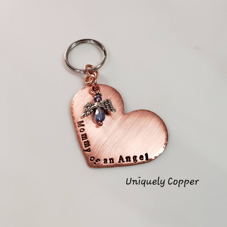 May include: Copper heart-shaped keychain with the text "Mommy of an Angel" engraved on it. A small silver angel charm with a blue crystal is attached to the heart. The keychain is attached to a silver key ring. The text "Uniquely Copper" is printed below the heart.