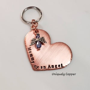 May include: Copper heart-shaped keychain with the words "Mommy of an Angel" engraved on it. A small angel charm with a blue crystal hangs from the top of the heart. The keychain is attached to a silver key ring.