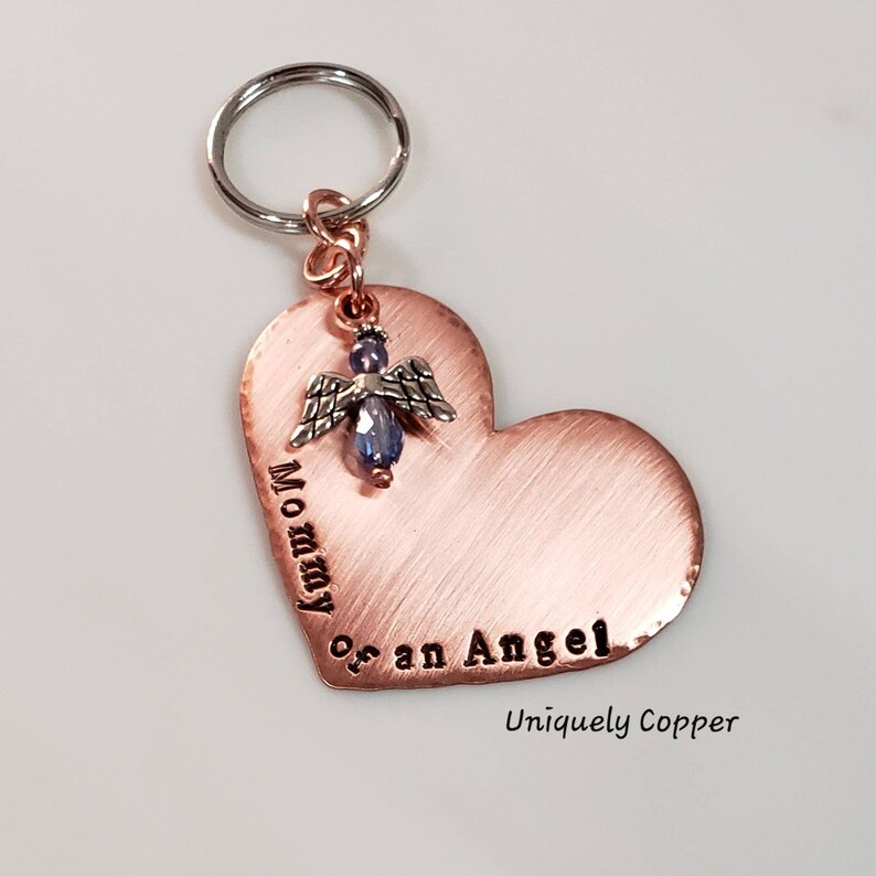 May include: Copper heart-shaped keychain with the words "Mommy of an Angel" engraved on it. A small angel charm with a blue crystal hangs from the top of the heart. The keychain is attached to a silver key ring.