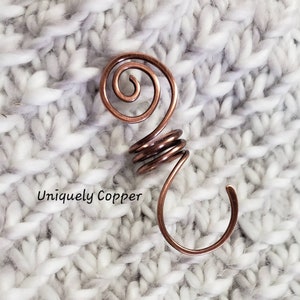 May include: A copper wire hook with a spiral design. The hook is on a white knitted surface. Uniquely Copper is written below the hook.