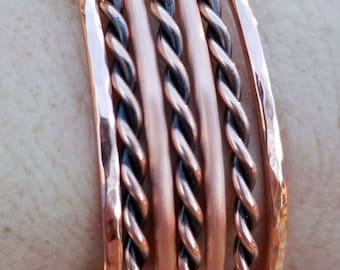May include: A set of five copper bracelets, each with a different texture. One bracelet is hammered, while the others are twisted or braided. The bracelets are stacked on top of each other.