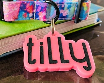 Puede incluir: Una etiqueta de perro personalizada de color rosa con el nombre "tilly" en letras negras. La etiqueta está unida a un collar de perro colorido con un patrón de tie-dye.