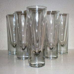 May include: A set of six clear, slender shot glasses. Each glass has a flared top and a rounded, heart-shaped base. The glasses are arranged in a semi-circle, showcasing their elegant design. They are made of transparent glass.