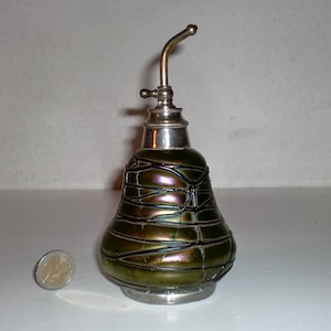 Puede incluir: Una botella de aceite de vidrio verde oliva con una boquilla y base de metal plateado. La botella tiene forma de pera con una superficie texturizada e iridiscente. Una moneda de euro está al lado de la botella para comparar el tamaño.