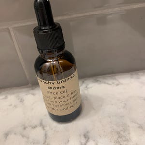 Op de afbeelding: Een bruine glazen fles Crunchy Granola Mama Face Oil met een zwarte druppelaar. Het etiket geeft instructies voor gebruik. De fles staat op een wit en grijs marmeren oppervlak.