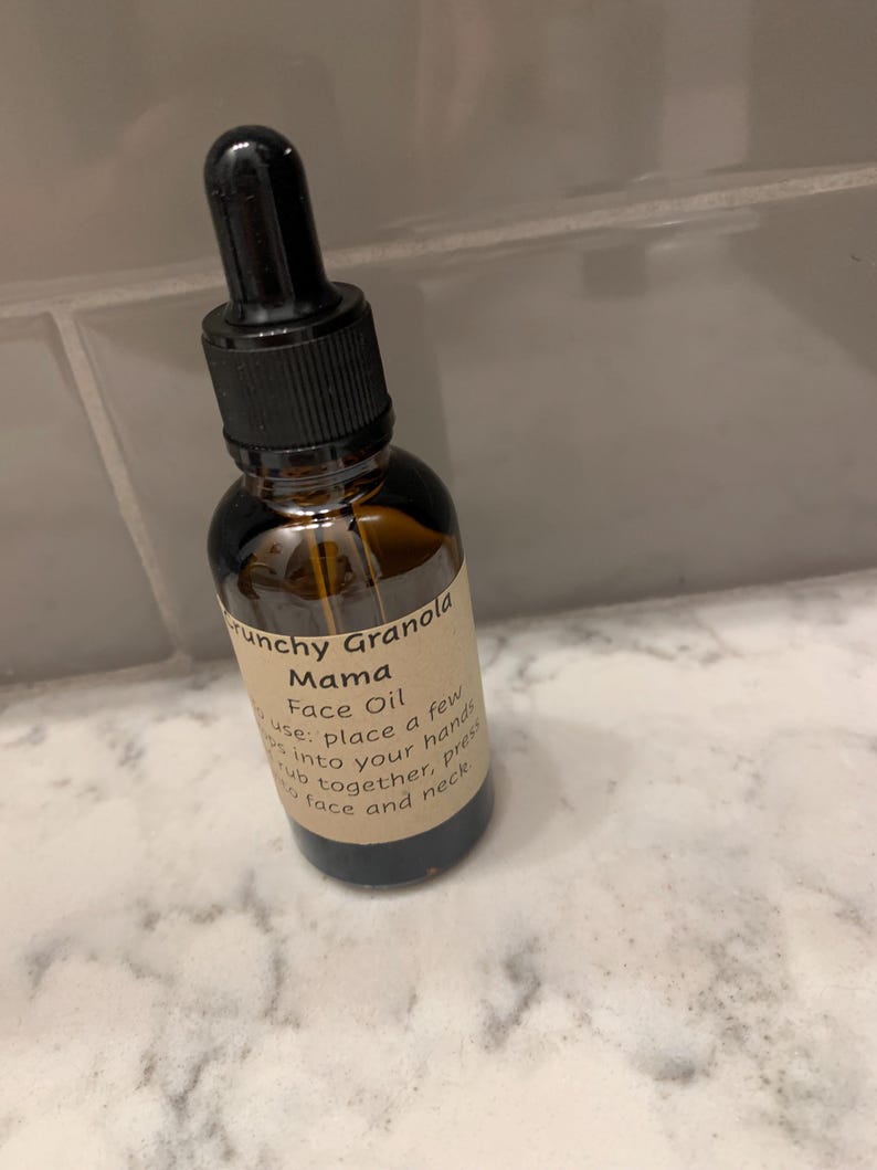Op de afbeelding: Een bruine glazen fles Crunchy Granola Mama Face Oil met een zwarte druppelaar. Het etiket geeft instructies voor gebruik. De fles staat op een wit en grijs marmeren oppervlak.