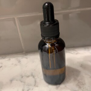 Op de afbeelding: Een donker amberkleurige glazen fles met een zwarte druppelaar. De fles heeft een zwarte dop en een heldere glazen body. De fles staat op een wit marmeren oppervlak.