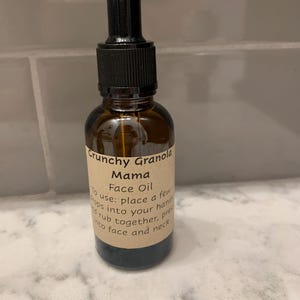 Op de afbeelding: Een bruine glazen fles Crunchy Granola Mama Face Oil met een zwarte druppelaar. Het etiket bevat instructies: "doe een paar druppels in je handen, wrijf samen, druk in gezicht en nek."