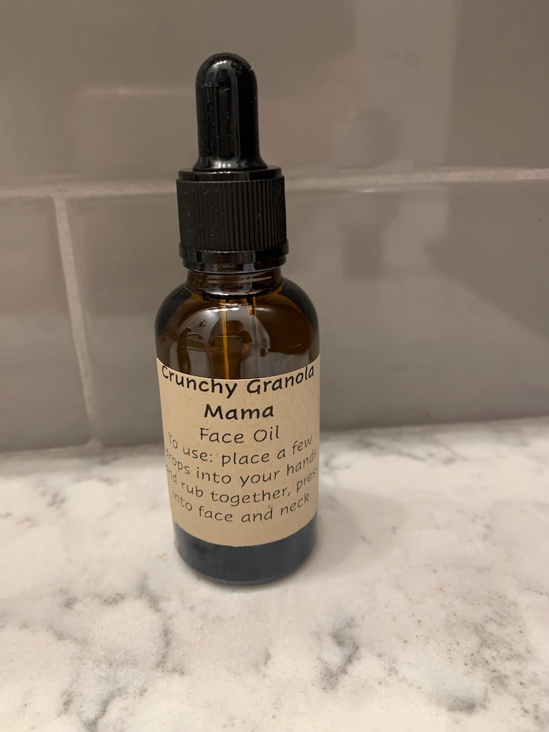 Op de afbeelding: Een bruine glazen fles Crunchy Granola Mama Face Oil met een zwarte druppelaar. Het etiket bevat instructies: "doe een paar druppels in je handen, wrijf samen, druk in gezicht en nek."