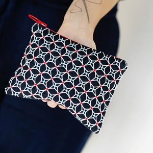 Peut inclure: Une pochette à motif géométrique noir et blanc avec des touches de rouge et une fermeture éclair rouge. La pochette est tenue dans la main d'une personne. De forme rectangulaire, elle est idéale pour petits objets.