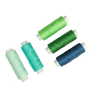 May include: An assortment of colorful thread spools in shades of green, teal, and blue. The spools are arranged diagonally against a white background. Each spool is cylindrical with a white core, and the thread is tightly wound.