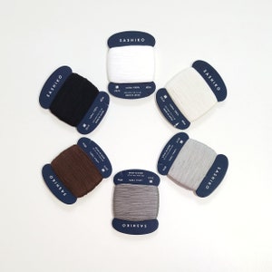 Puede incluir: Seis carretes de hilo de diferentes colores, incluyendo blanco, marrón, negro y gris. Cada carrete está etiquetado con "SASHIKO" y tiene detalles sobre el hilo, como el material, la longitud y el tamaño.