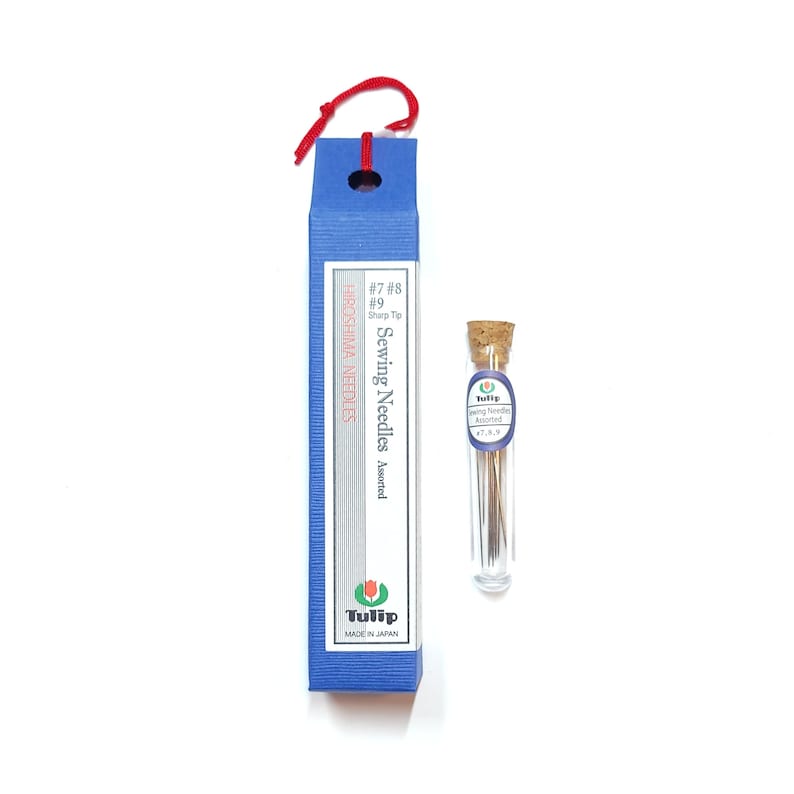 May include: A blue box with a red string handle contains a pack of Tulip brand sewing needles. The box is labelled "Hiroshima Needles" and "Sewing Needles Assorted". The needles are also in a clear glass tube with a cork top.