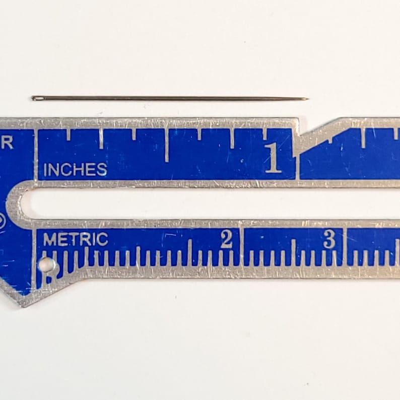 May include: A close-up of a silver ruler with blue markings. The ruler has both inches and metric measurements. A thin, silver needle is placed on top of the ruler, measuring approximately 1 inch in length.