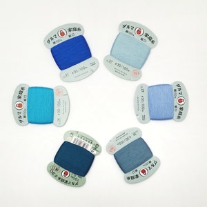 May include: Five spools of blue, teal, and light blue thread. Each spool is labeled "ダルマ 家庭糸" which translates to "Daruma Family Thread" and "綿100%" which translates to "100% Cotton". The spools are labeled with different numbers and lengths, such as "#30/100m".