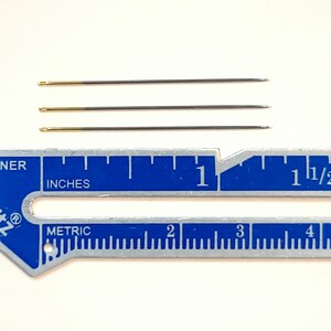 May include: Four gold-tipped sewing needles measuring approximately 2.5 cm in length, displayed against a blue ruler with inch and metric markings.