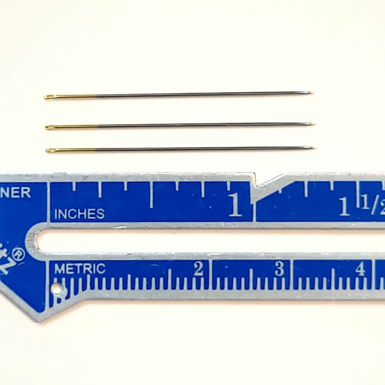 May include: Four gold-tipped sewing needles measuring approximately 2.5 cm in length, displayed against a blue ruler with inch and metric markings.
